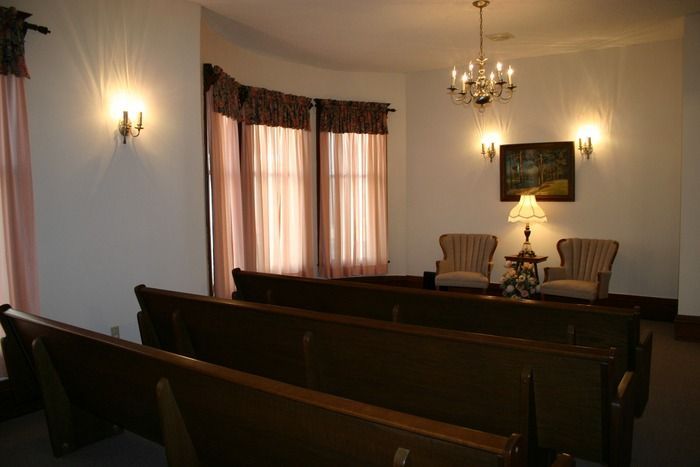 A formal viewing room with rows of wooden pews facing two chairs, a lamp, and a wall painting under a chandelier.