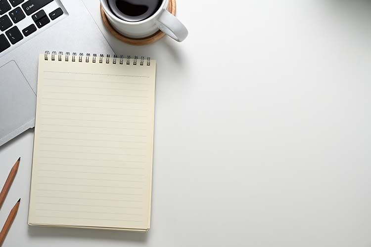 A top-down view of a white desk with a laptop, a cup of coffee, a blank spiral notebook, and two pencils.