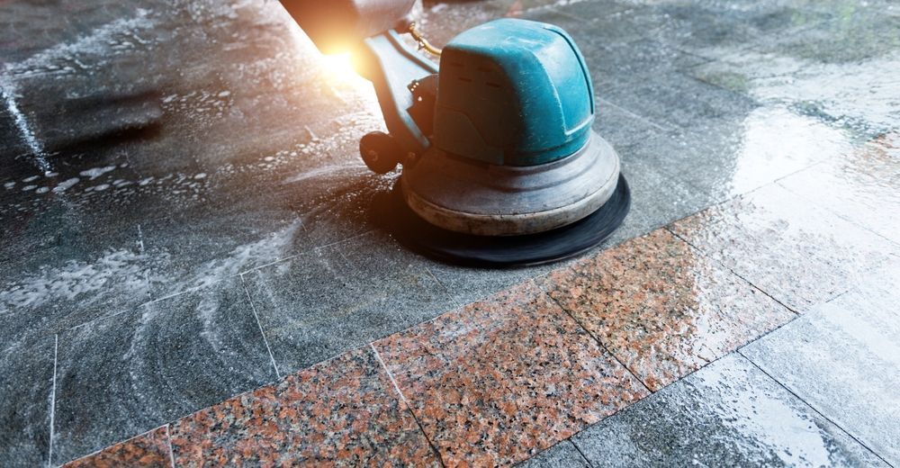 A floor buffer scrubs a wet, patterned granite floor with cleaning solution, shining light.