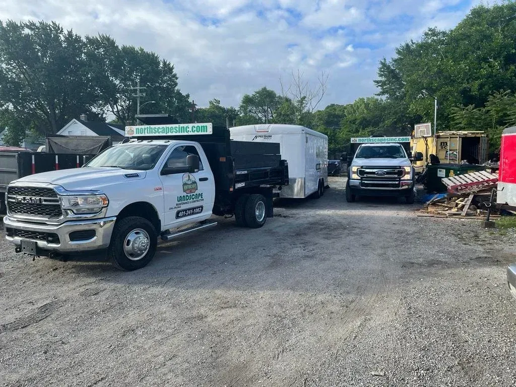 White work trucks and a utility trailer parked on a gravel lot beside trees under a cloudy sky