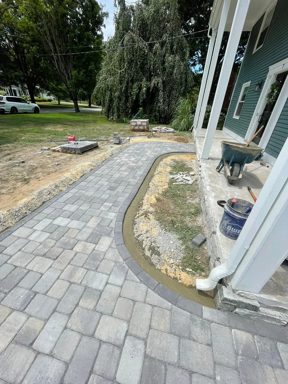 A newly installed paver walkway curves along the porch of a teal house, with construction tools nearby on a lawn.