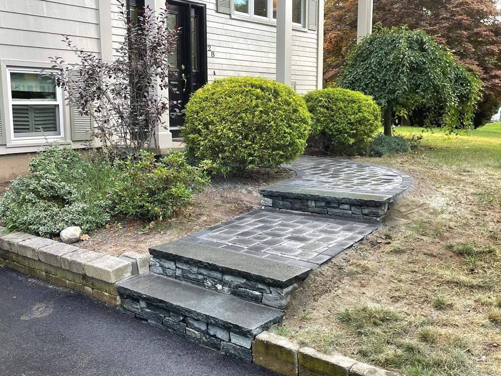 A stone-paved walkway with two steps leading from a dark asphalt driveway to the front entrance of a house.