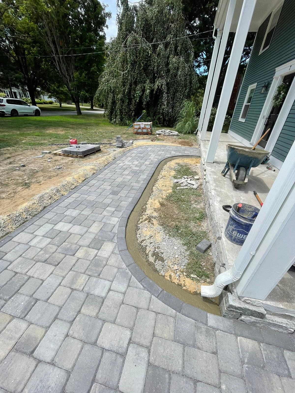 A new paver walkway curves along the porch of a house under construction, with a wheelbarrow and bucket nearby.