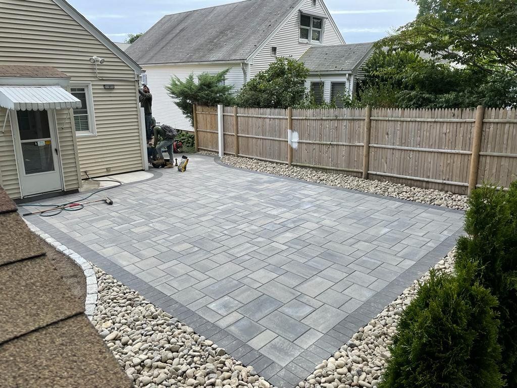 A newly installed grey paver patio with a dark border, surrounded by light-colored decorative stones in a backyard.
