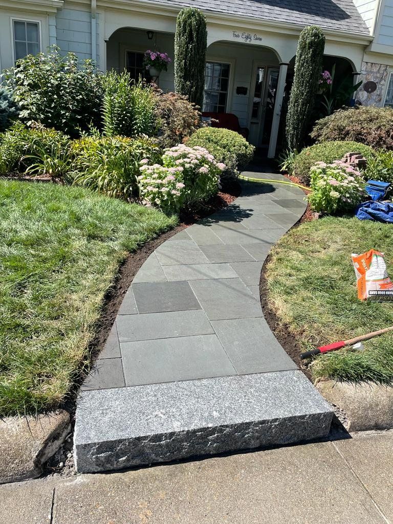 A bluestone walkway leads to a house entrance, featuring a wide granite step at the start and manicured garden beds.