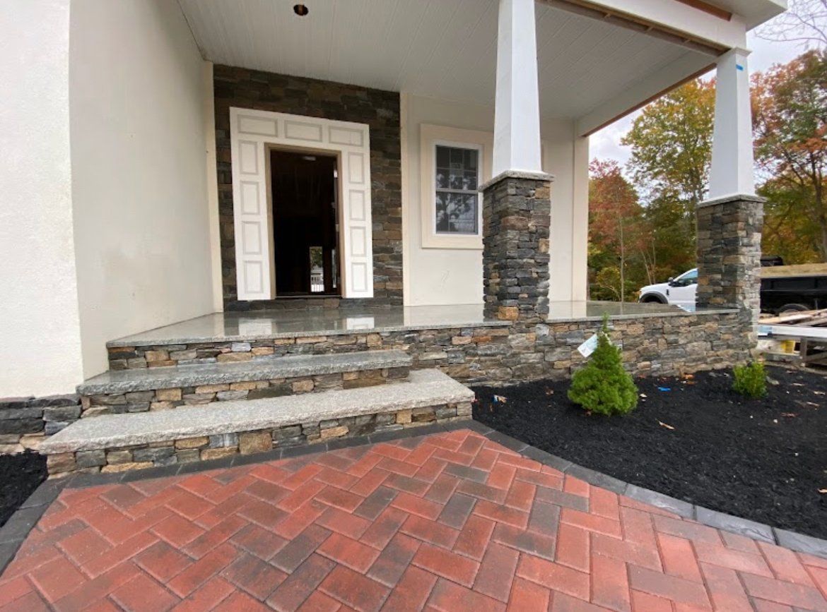 A front porch with stone-veneer steps, a herringbone-patterned red brick walkway, and two white pillars.