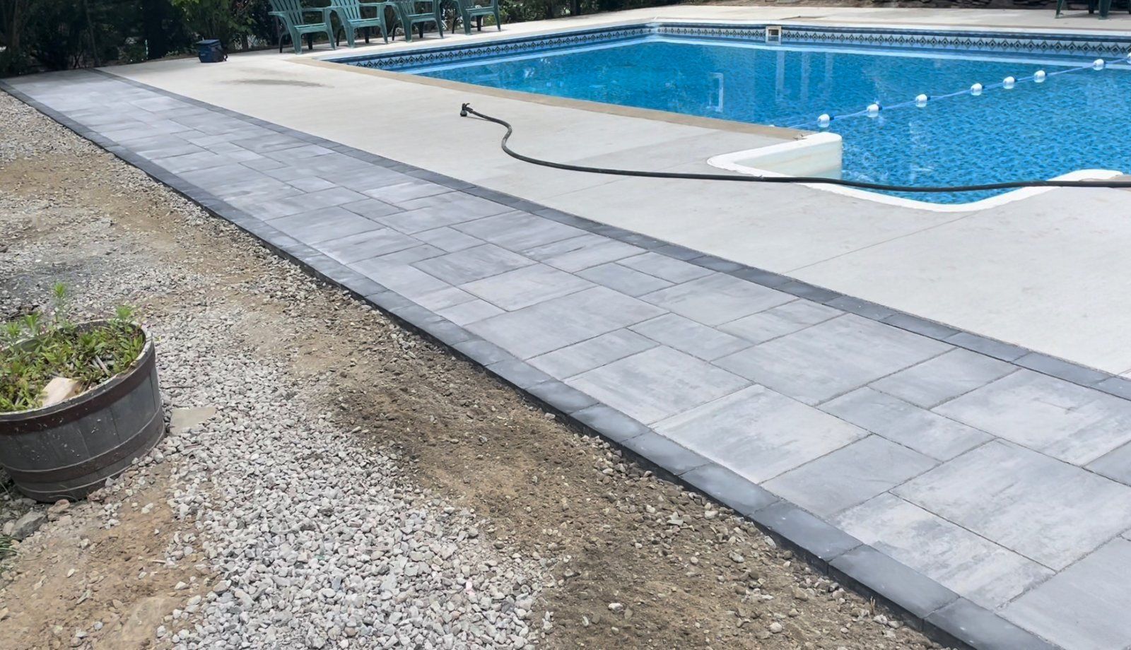 A gray paved walkway borders a concrete pool deck and a blue swimming pool, next to a patch of gravel and a planter.