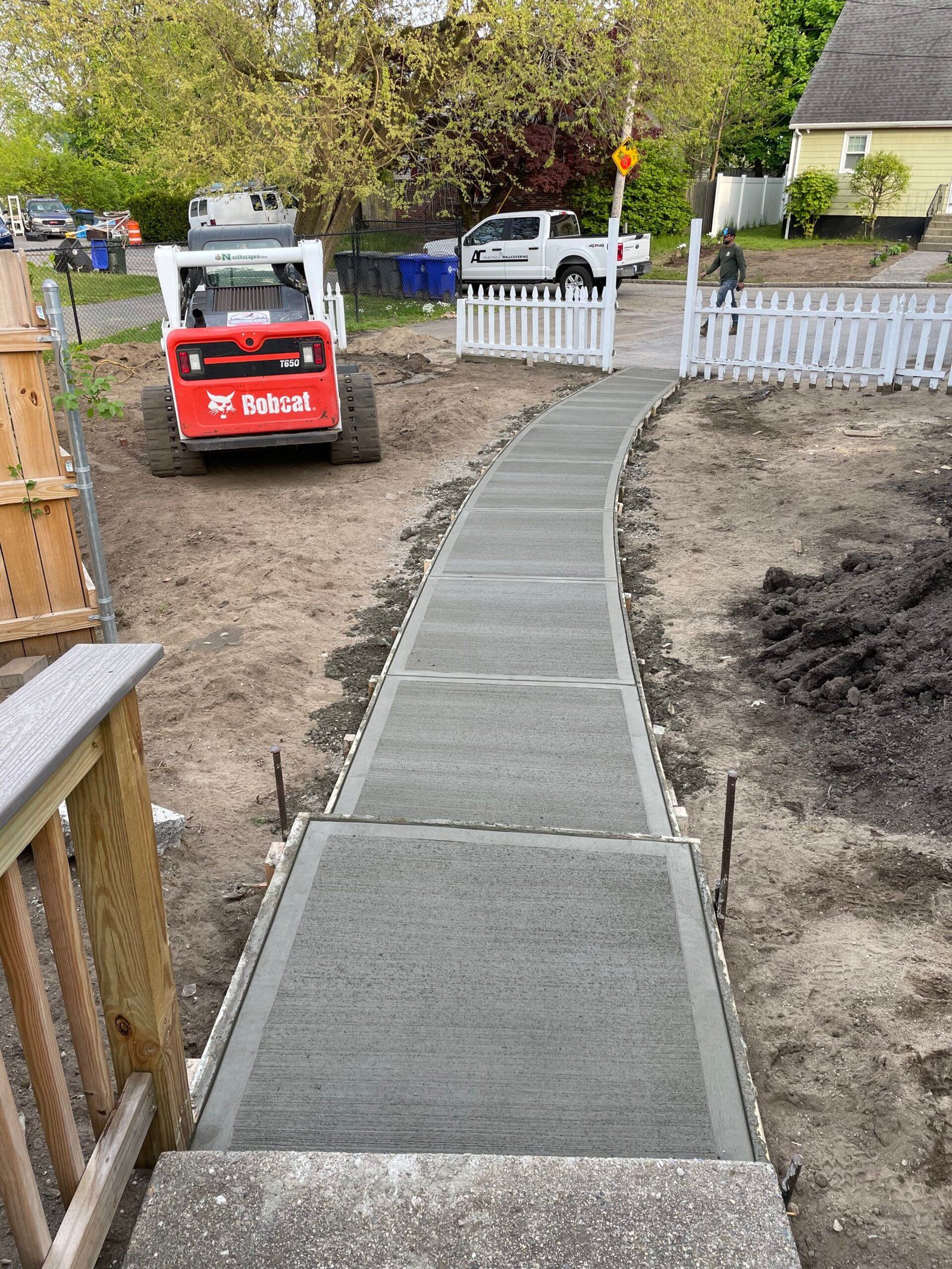 A freshly poured concrete sidewalk leads to a white picket fence, with a red Bobcat skid steer parked on the left.