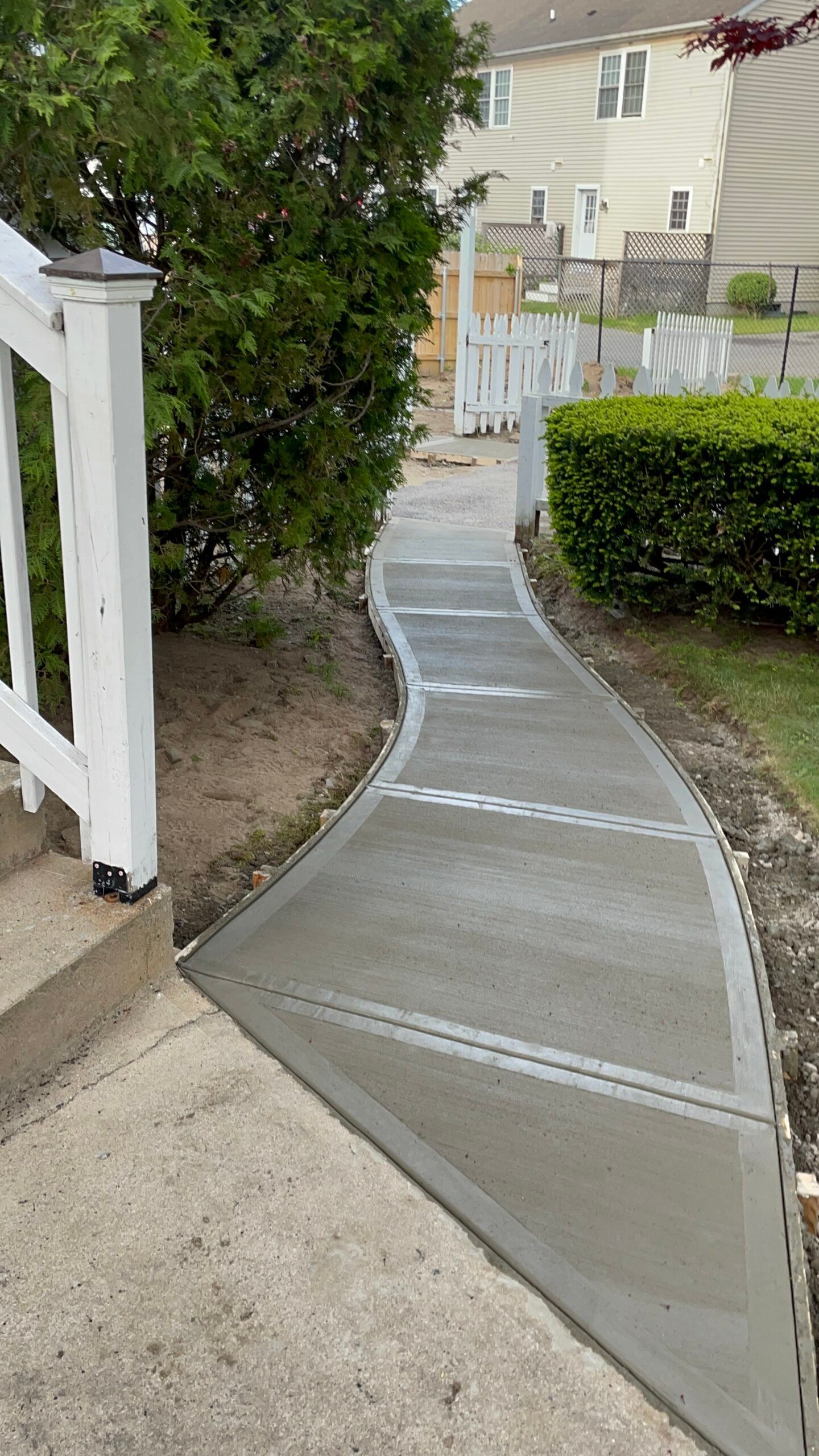 A newly poured concrete walkway curves from a set of stairs through a residential lawn toward a white fence.