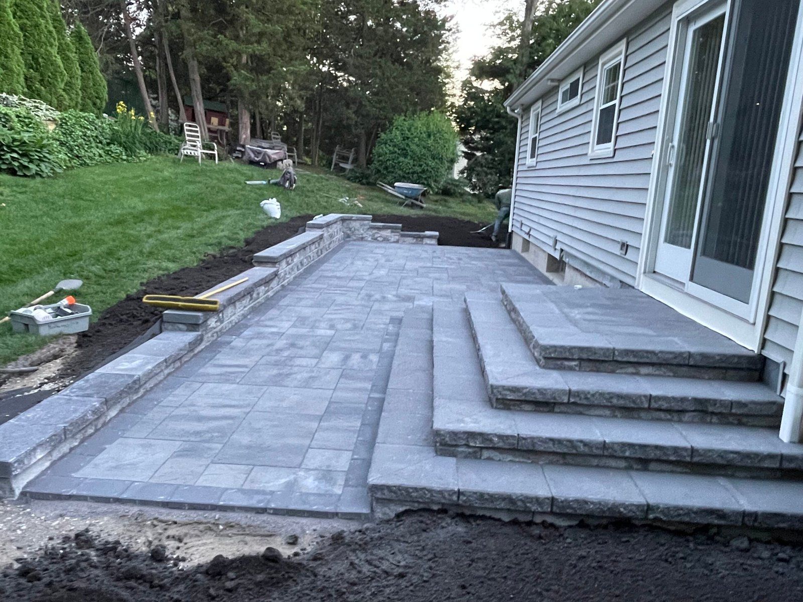 A gray stone patio under construction outside a house, featuring newly installed steps leading to a sliding glass door.