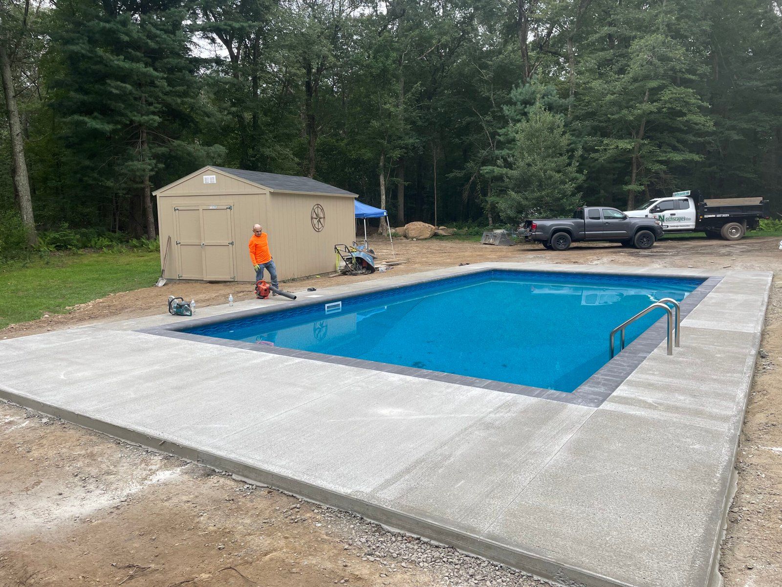 An outdoor rectangular swimming pool with new concrete decking, a shed, and trucks in a wooded backyard setting.