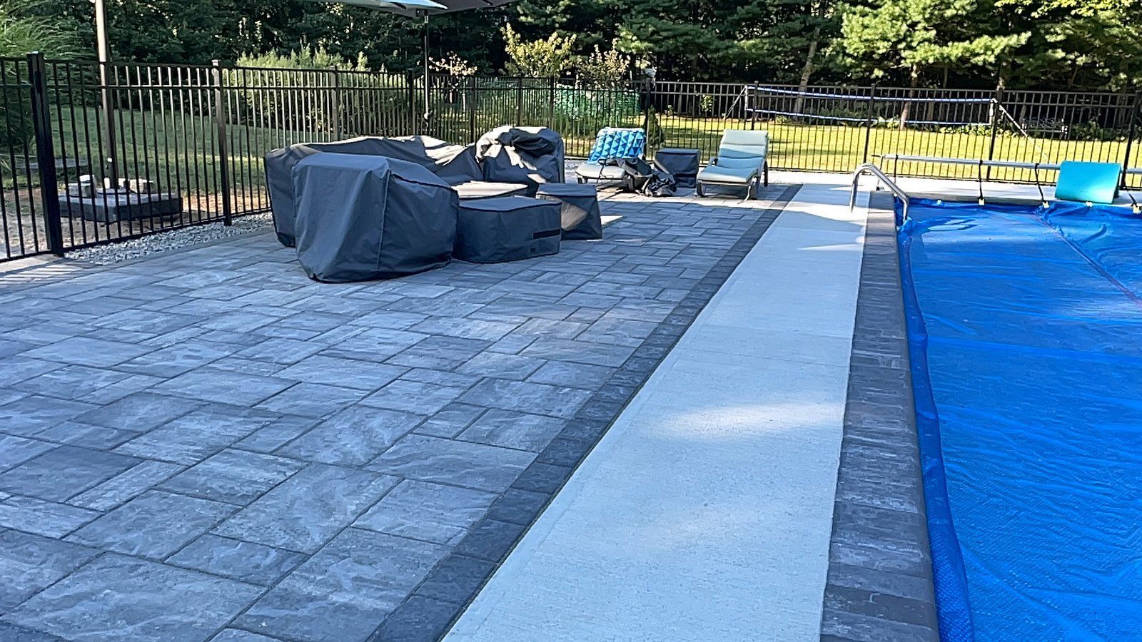 A backyard pool deck with gray stone pavers, a white concrete border, covered furniture, and a blue pool cover.