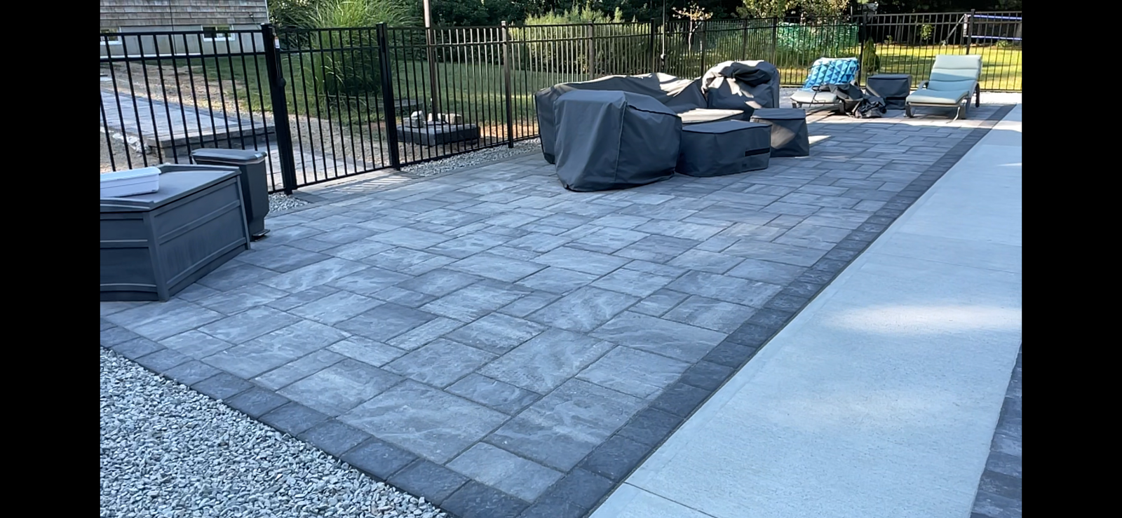 Gray paver patio with a dark border, outdoor furniture covered in gray fabric, and a nearby light-colored walkway.