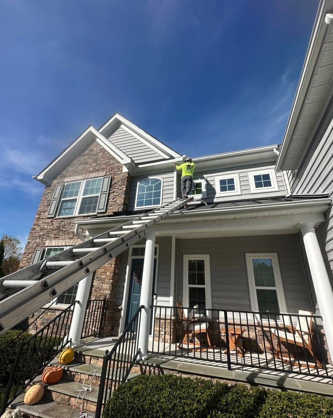 Worker on ladder repairing roof of stone house with porch under clear blue sky