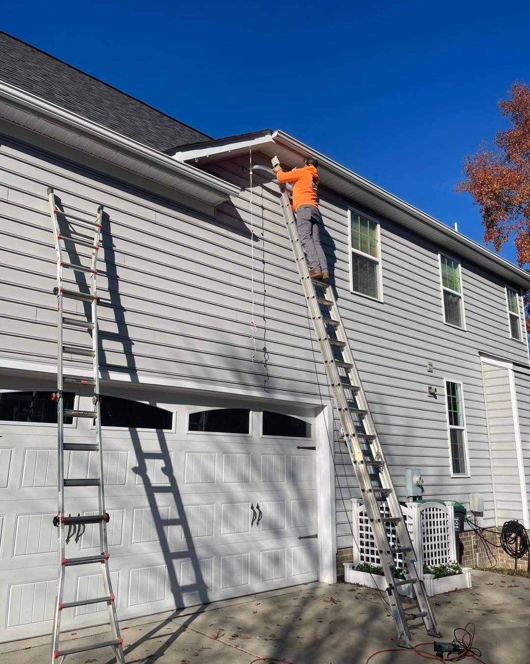 Worker on a ladder painting white siding on a gray house under a clear blue sky.