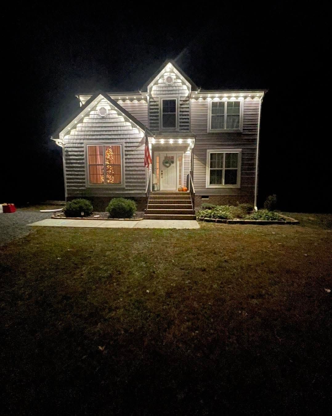 Well-lit house at night with white siding, lit windows, front porch, and a dark yard