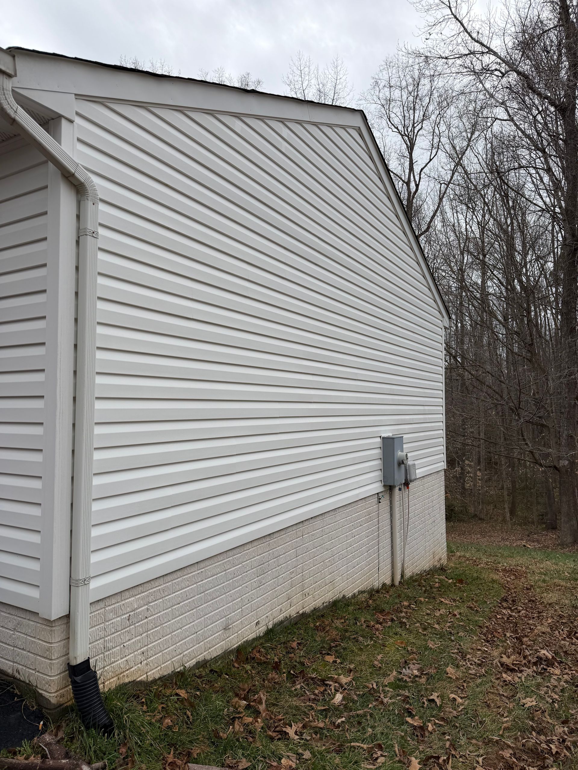 White vinyl-sided house exterior with a side wall, downspout, and leaf-covered yard under cloudy sky
