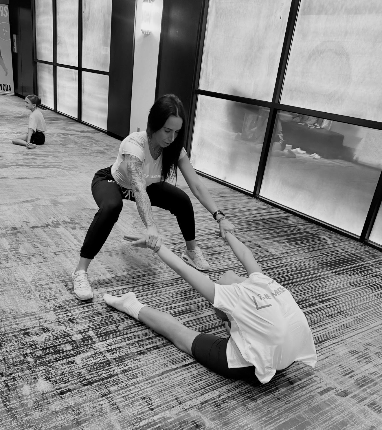 Woman assisting a person in a seated split stretch. Training on a patterned carpet.