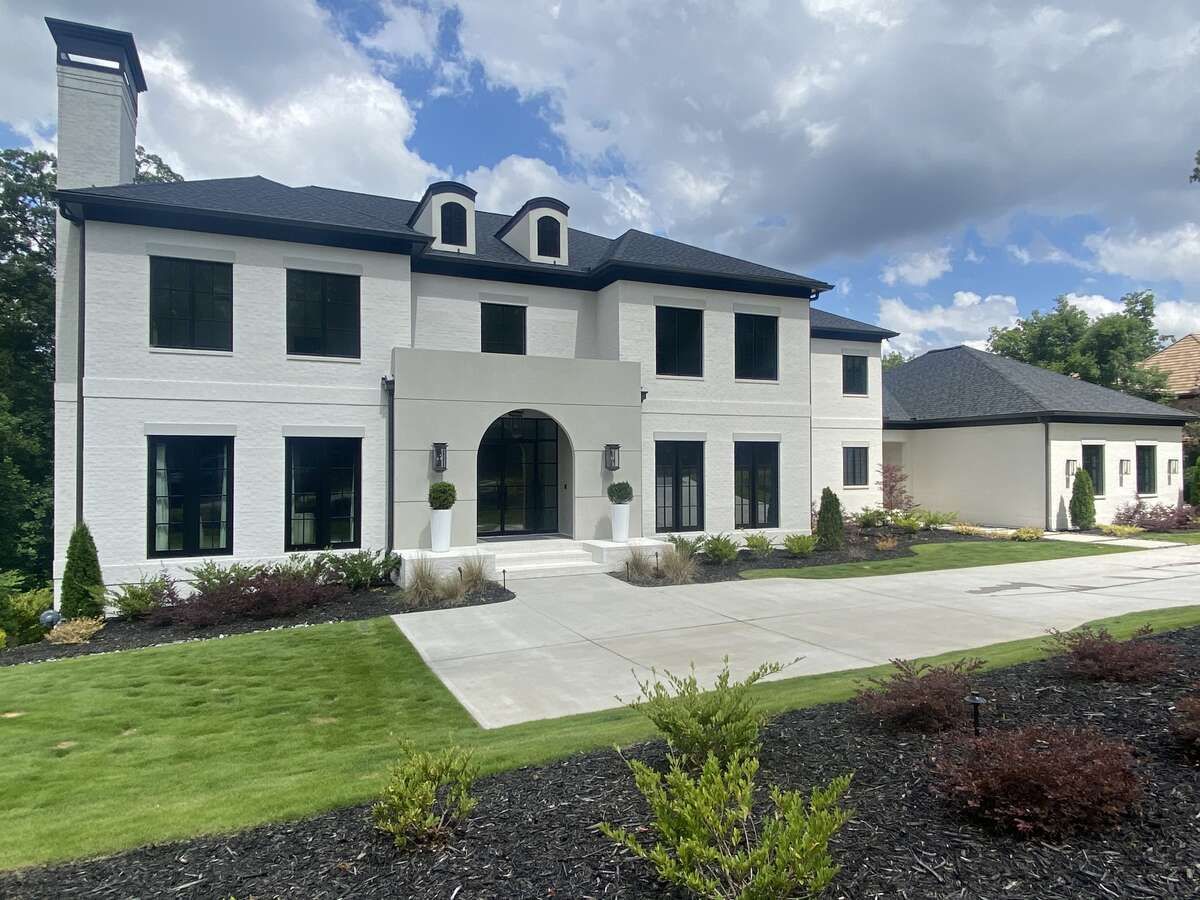 White two-story house with black windows, arched entryway, and dark roof. Driveway leads to the house, with lawn and dark mulch.