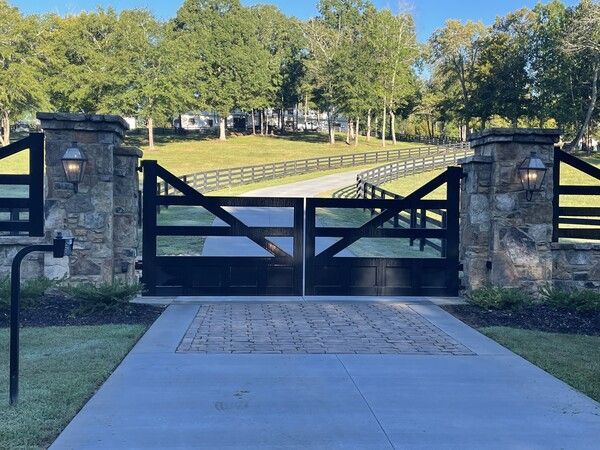 Black gates on brick columns, leading to a long driveway and fenced pasture under a sunny sky.