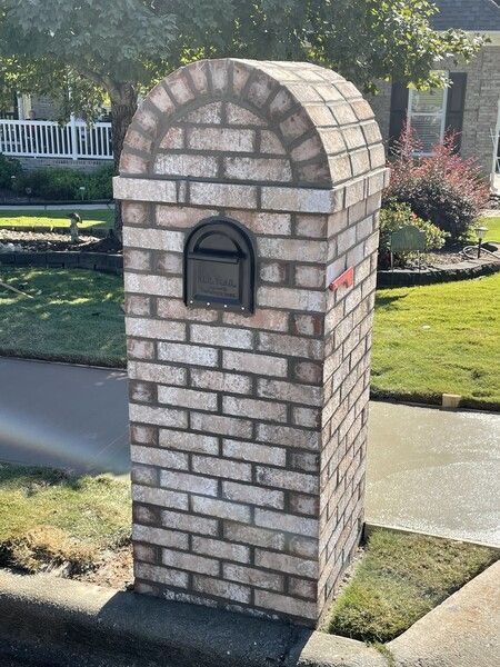 Brick mailbox with arched top and black metal mailbox against a grassy yard and street.