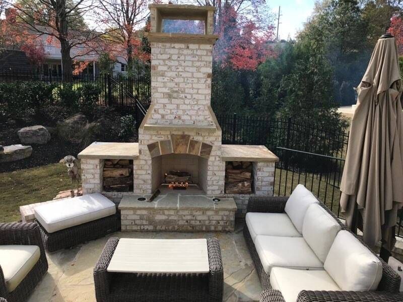 Outdoor stone fireplace with seating area. Firewood, trees, and patio furniture visible.