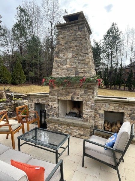 Outdoor stone fireplace with seating area; greenery and trees in the background.