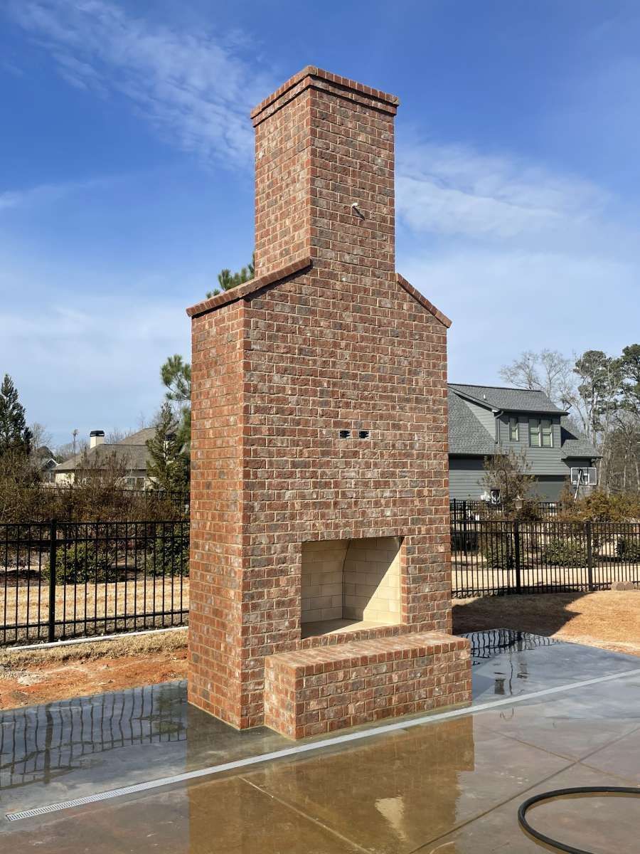 Brick outdoor fireplace with chimney against a blue sky, near houses and a fence.