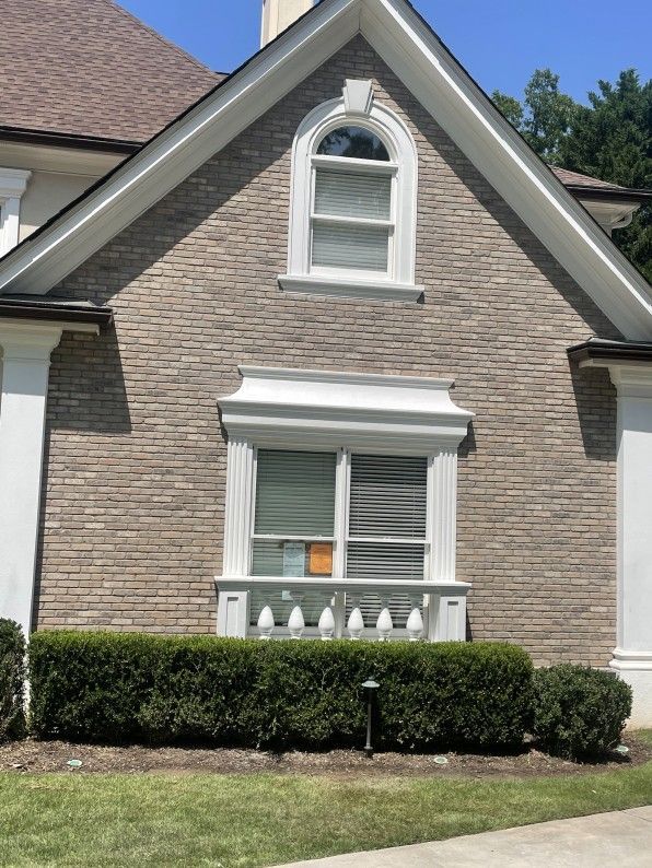 Light brick exterior of a house with white trim around two windows and a neatly trimmed green hedge.