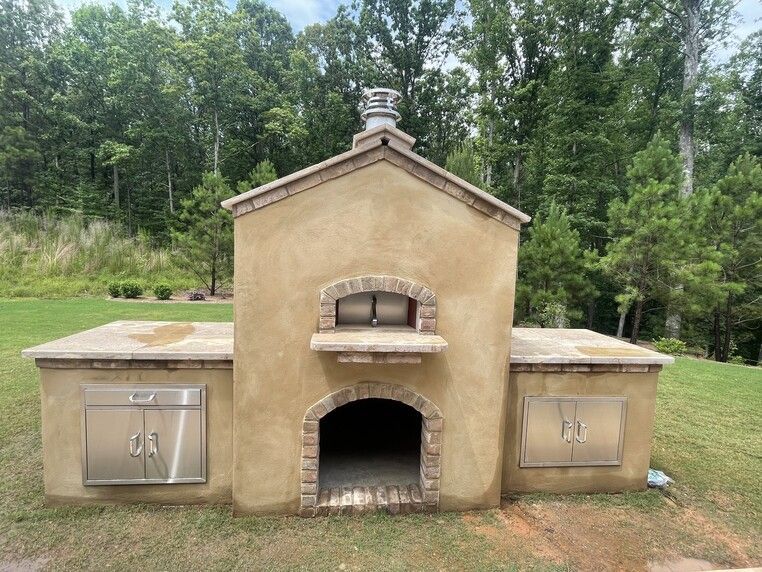 Outdoor brick pizza oven with chimney, tan stucco, and stainless steel doors. Surrounded by grass and trees.