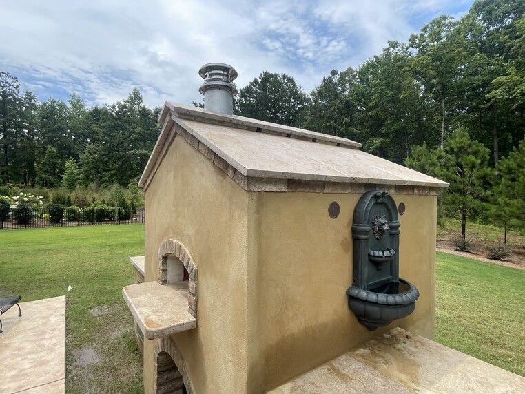 Outdoor pizza oven with a water fountain, tan stucco, stone trim, and a green yard with trees.