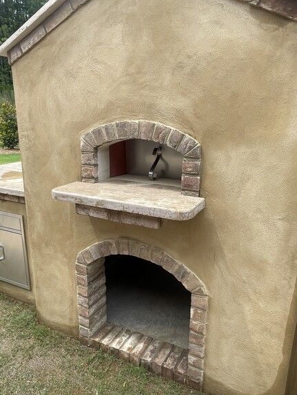 Outdoor pizza oven with brick-lined openings, beige stucco, and a stone shelf.