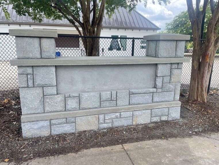 Stone sign with a blank center, capped with gray pillars, by a chain link fence.