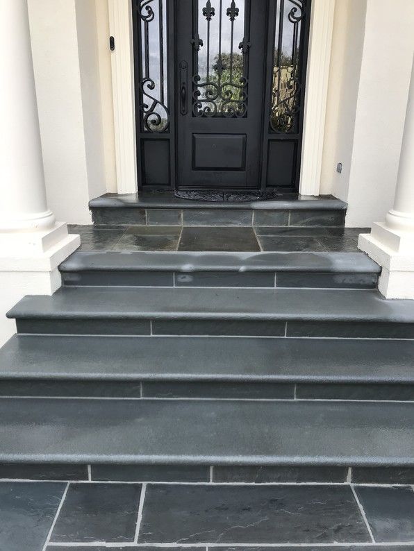 Dark gray stone steps leading to a black door with decorative ironwork, flanked by white columns.