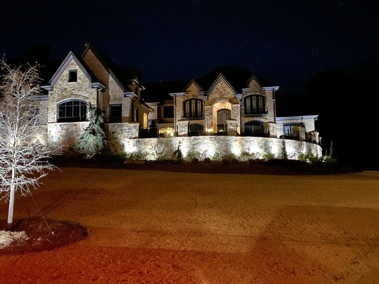 Nighttime view of a large stone house illuminated by spotlights along its foundation; dark sky.