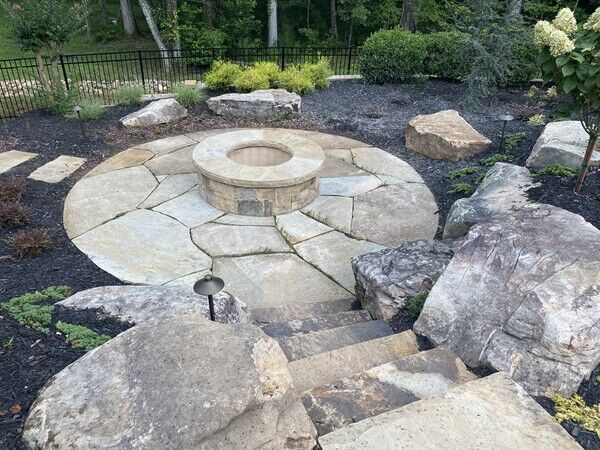 Stone fire pit in a backyard with stone steps and large rocks.