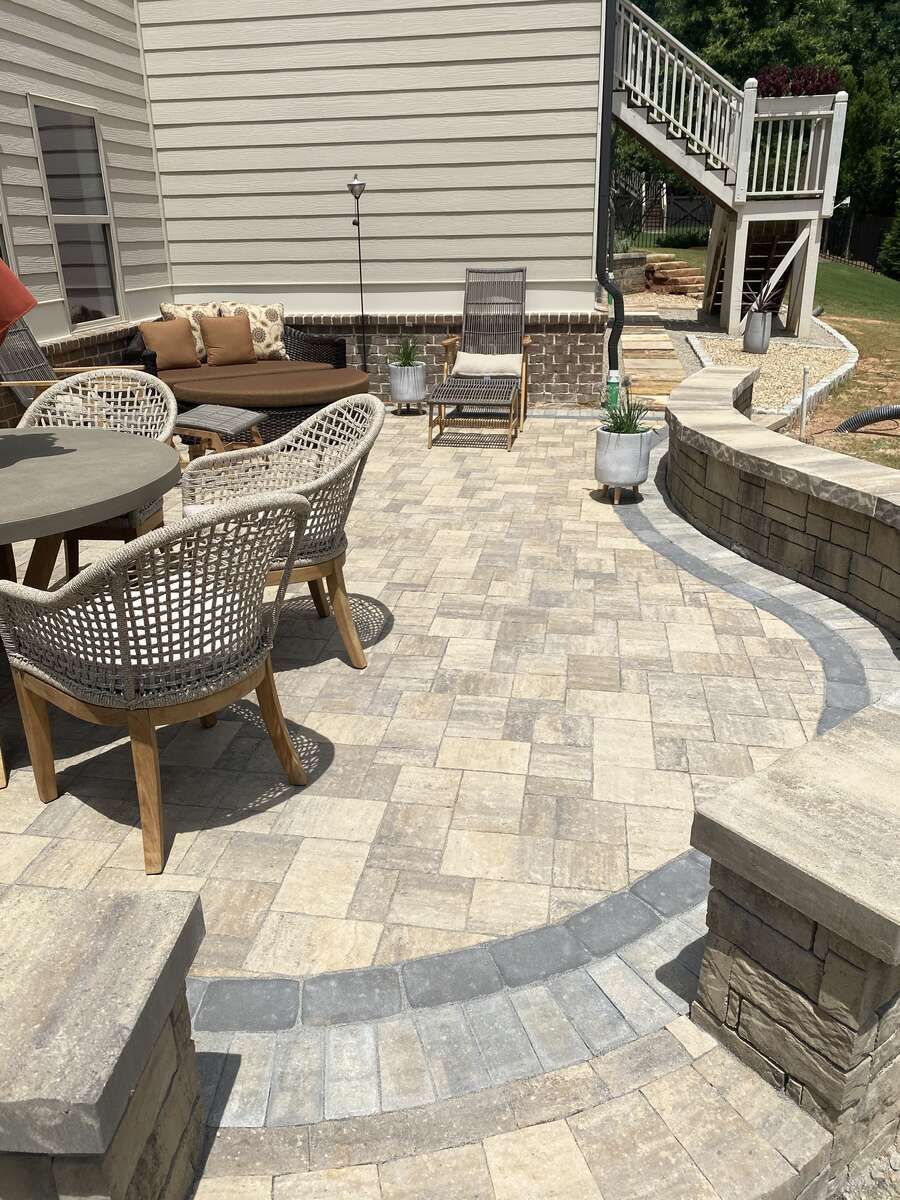 Patio with tan pavers, seating, and a retaining wall near a house.
