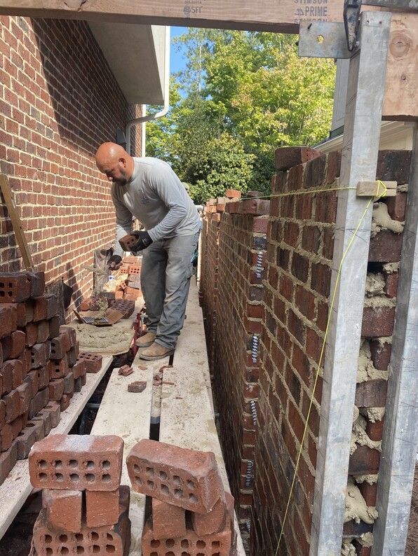 Bricklayer building a brick wall between supports. Bricks are red-brown. He is using a trowel.
