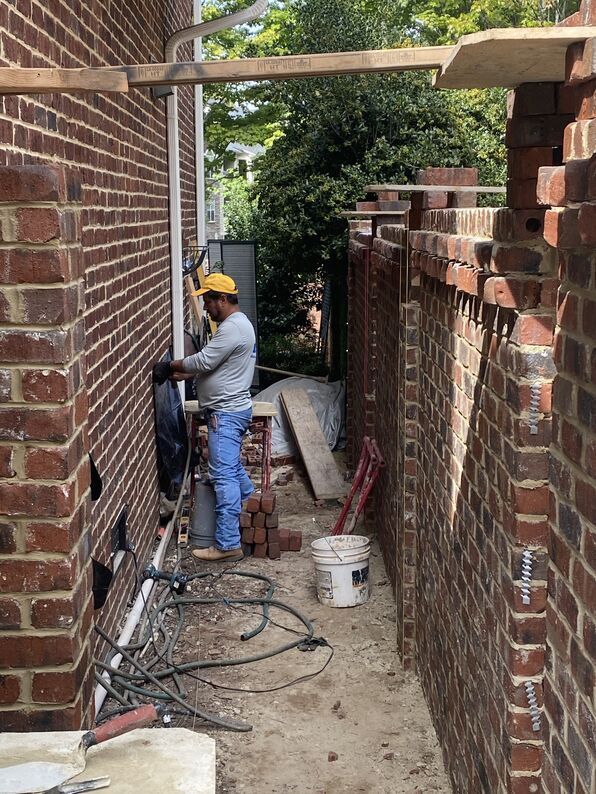 Construction worker in a narrow brick alleyway, using tools near a partially built brick wall. Yellow hard hat, daylight.
