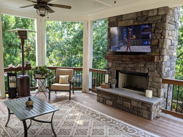 Outdoor living room with stone fireplace, TV, and seating on a deck, overlooking trees.