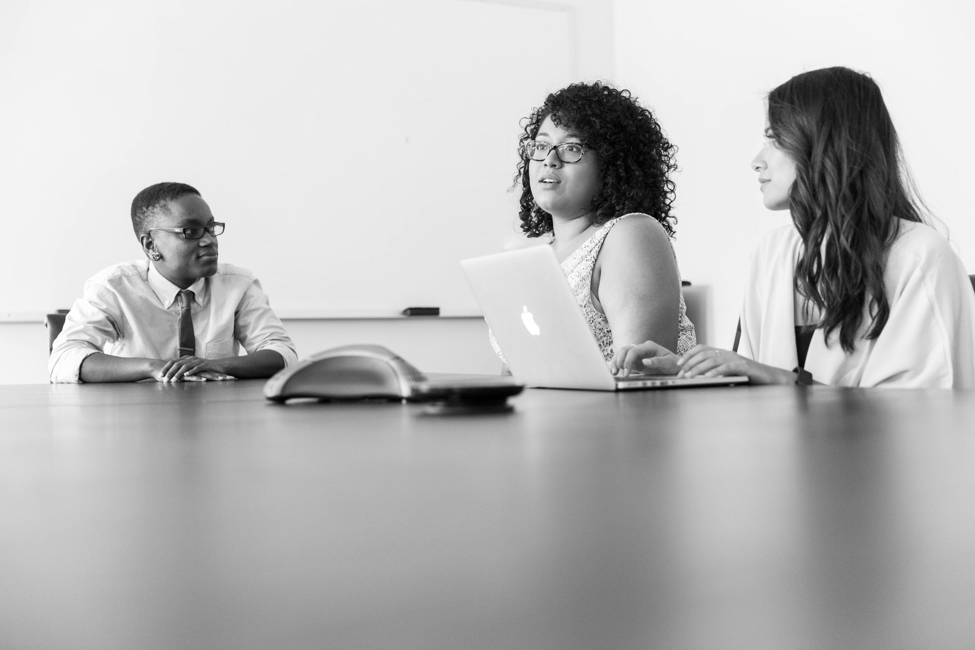 Three people in a meeting, one using a laptop, discussing at a table in a bright room.