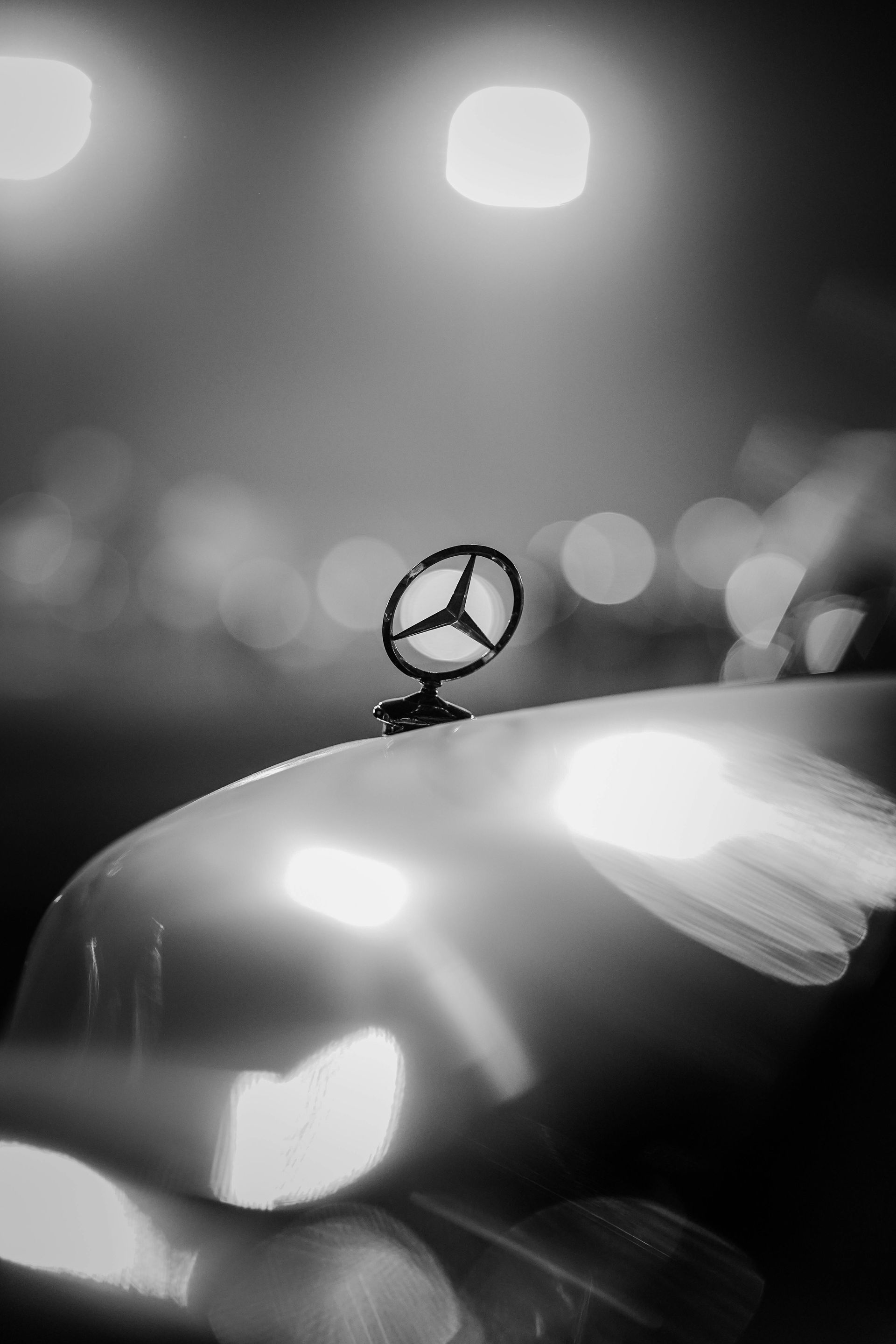 Mercedes hood ornament, car headlights, and blurred city lights in black and white.