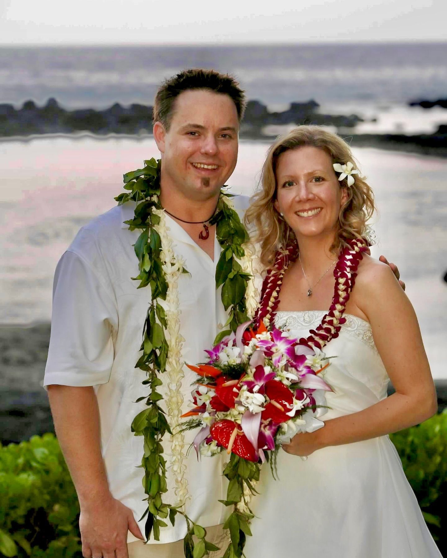 Couple in white wedding attire with floral leis standing by the ocean at sunset
