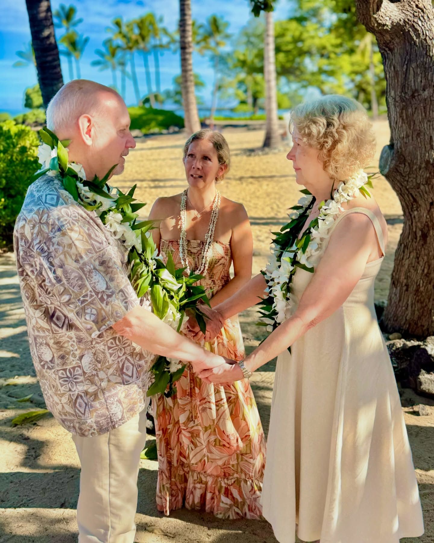 Three people hold hands in a tropical outdoor wedding ceremony under palm trees.
