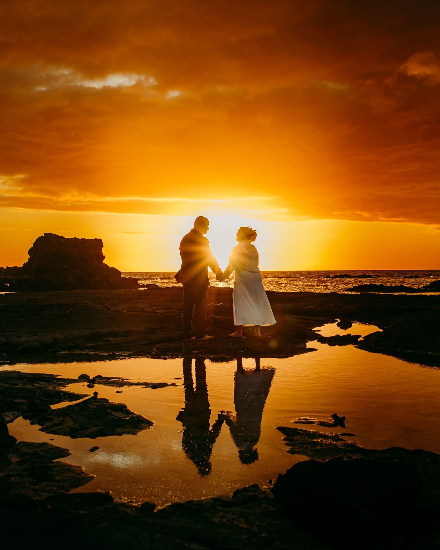 Silhouetted couple standing on a rocky shore at sunset, reflected in a tide pool under a golden sky