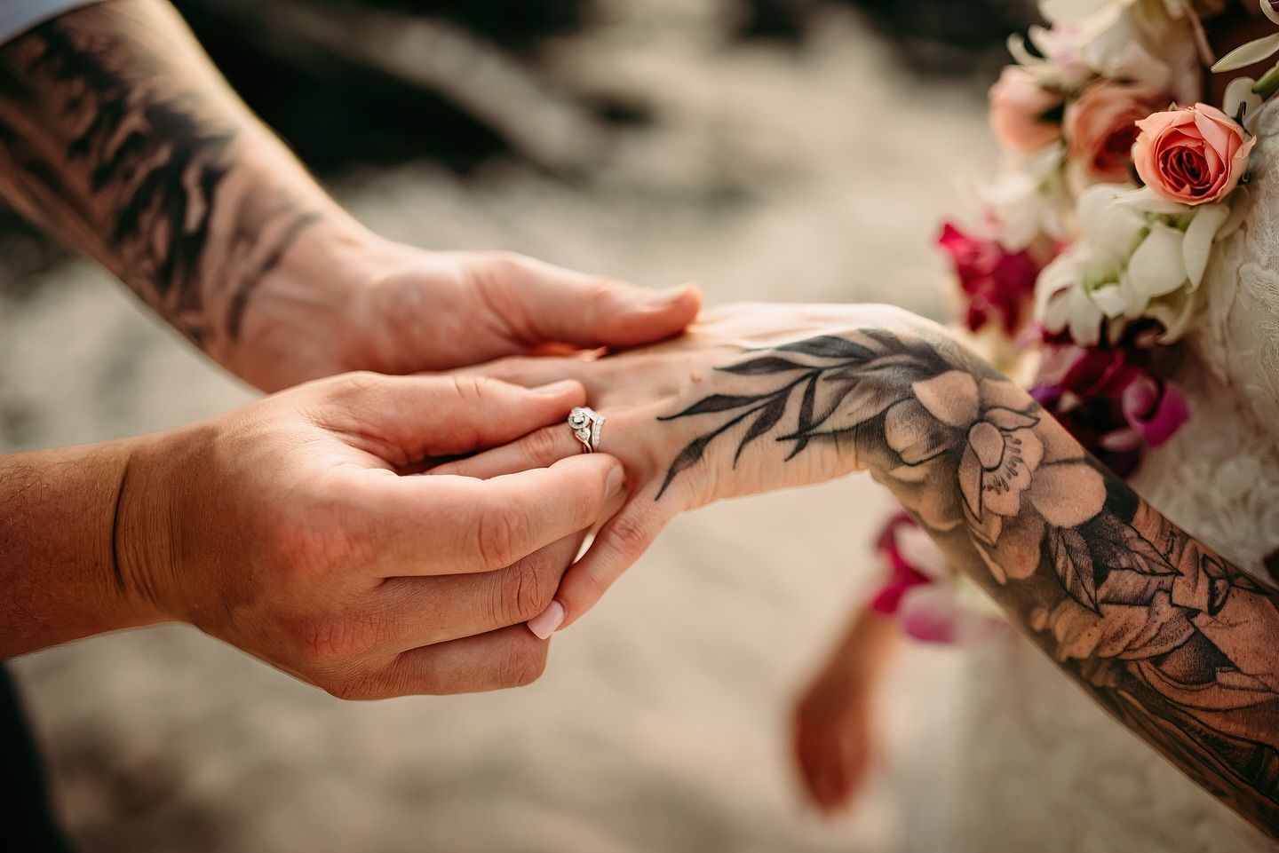 Two tattooed hands holding, one placing a ring on the other, with flowers in the background