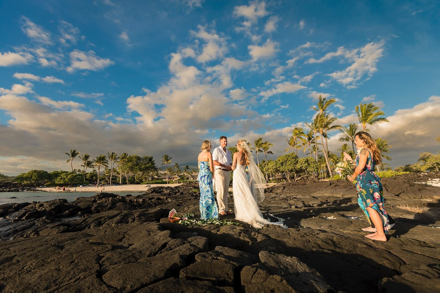 Simple Kona Beach Weddings wedding in action, the bride wearing white and a long veil, the groom also in white, the officiant, Becky Ringler, wearing blue. They stand on lava on a Kailua-Kona Beach, with a blue sky and palm trees in the background.
