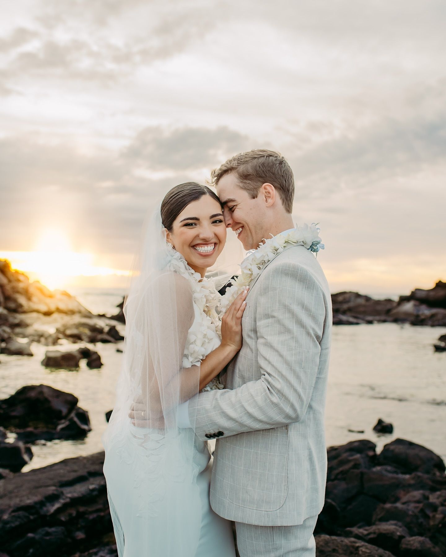 Bride and groom embracing on a rocky beach at sunset, smiling in wedding attire