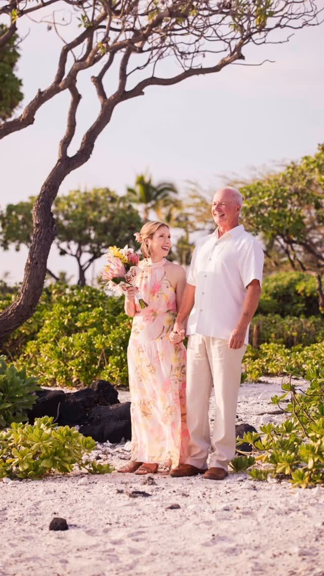 Two people stand in a tropical garden, holding a bouquet and smiling near palm trees.