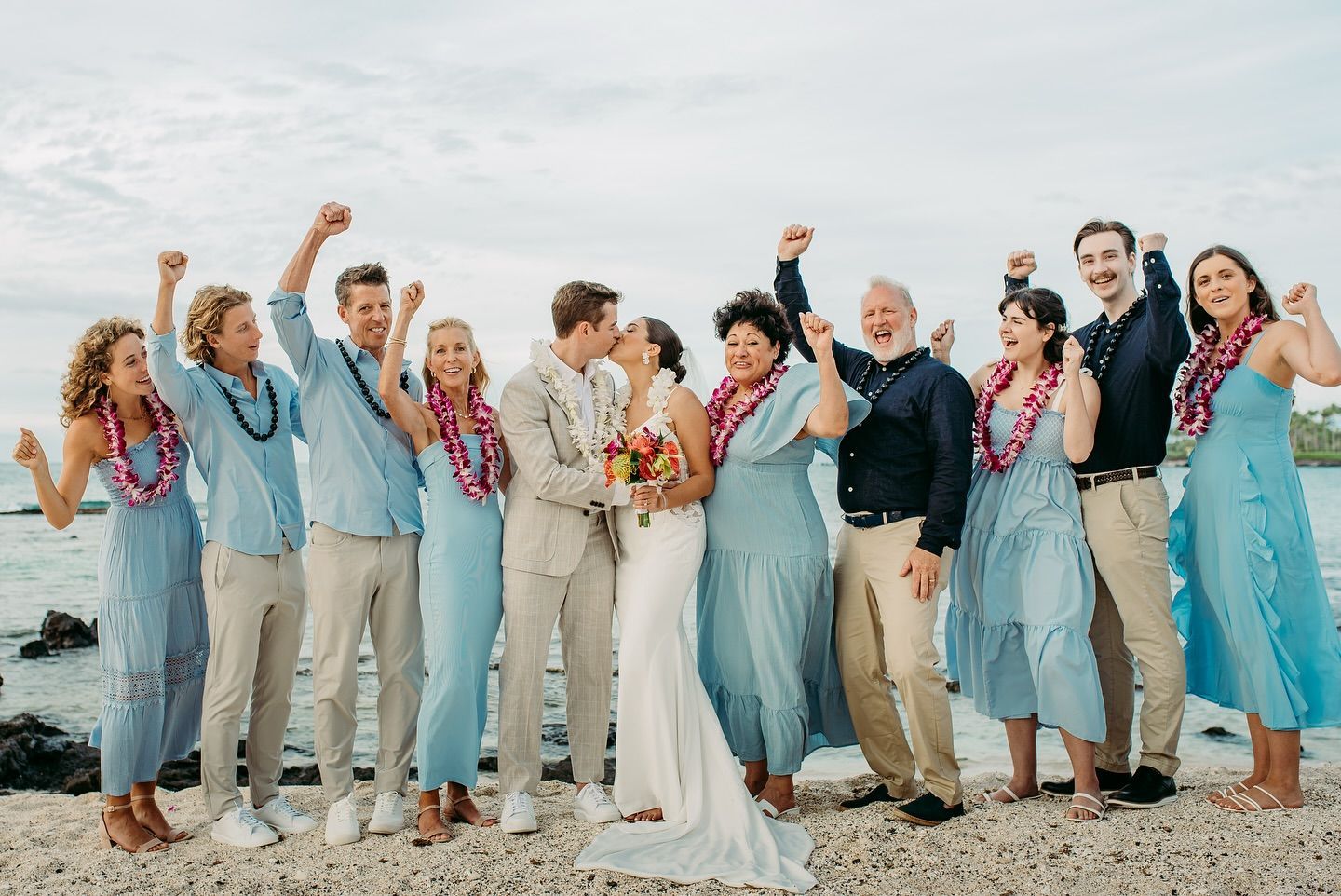 Wedding group celebrates on beach; bride and groom kiss. Light blue outfits, raised arms, leis.